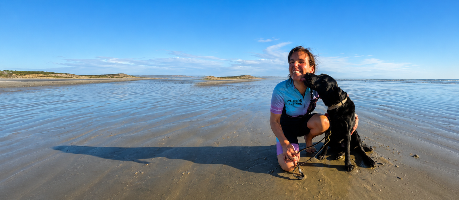 With the beach in the background, Hannah wears her Chasing Dreams cycling kit while her black guide dog Dara licks her face.