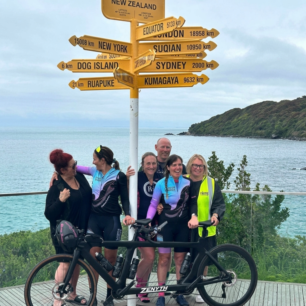 Hannah and her support crew standing beside her tandem bike and the Stirling Point sign in Bluff.