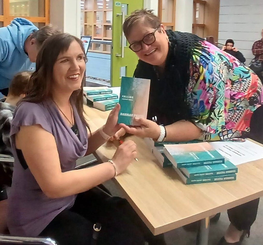 Hannah sitting at a table signing her book, with an older woman standing beside her. Both are smiling at the camera.