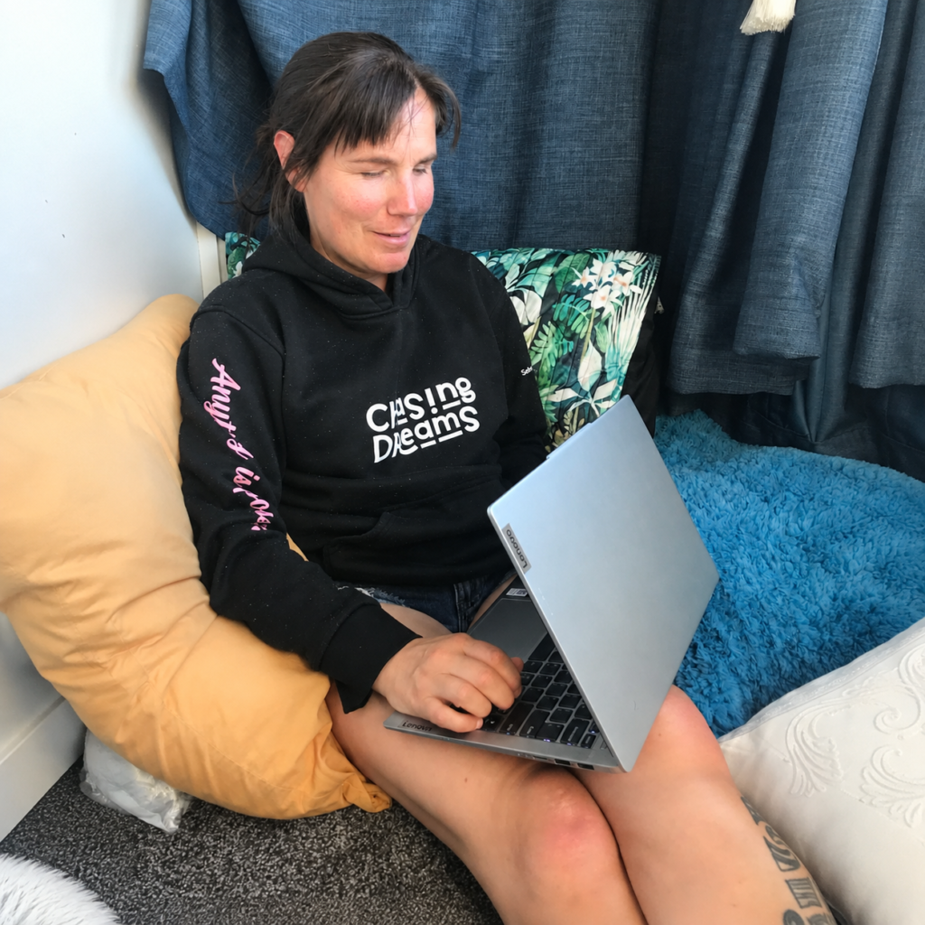 Hannah sitting in a corner surrounded by pillows, wearing her Chasing Dreams hoodie with a laptop on her lap.