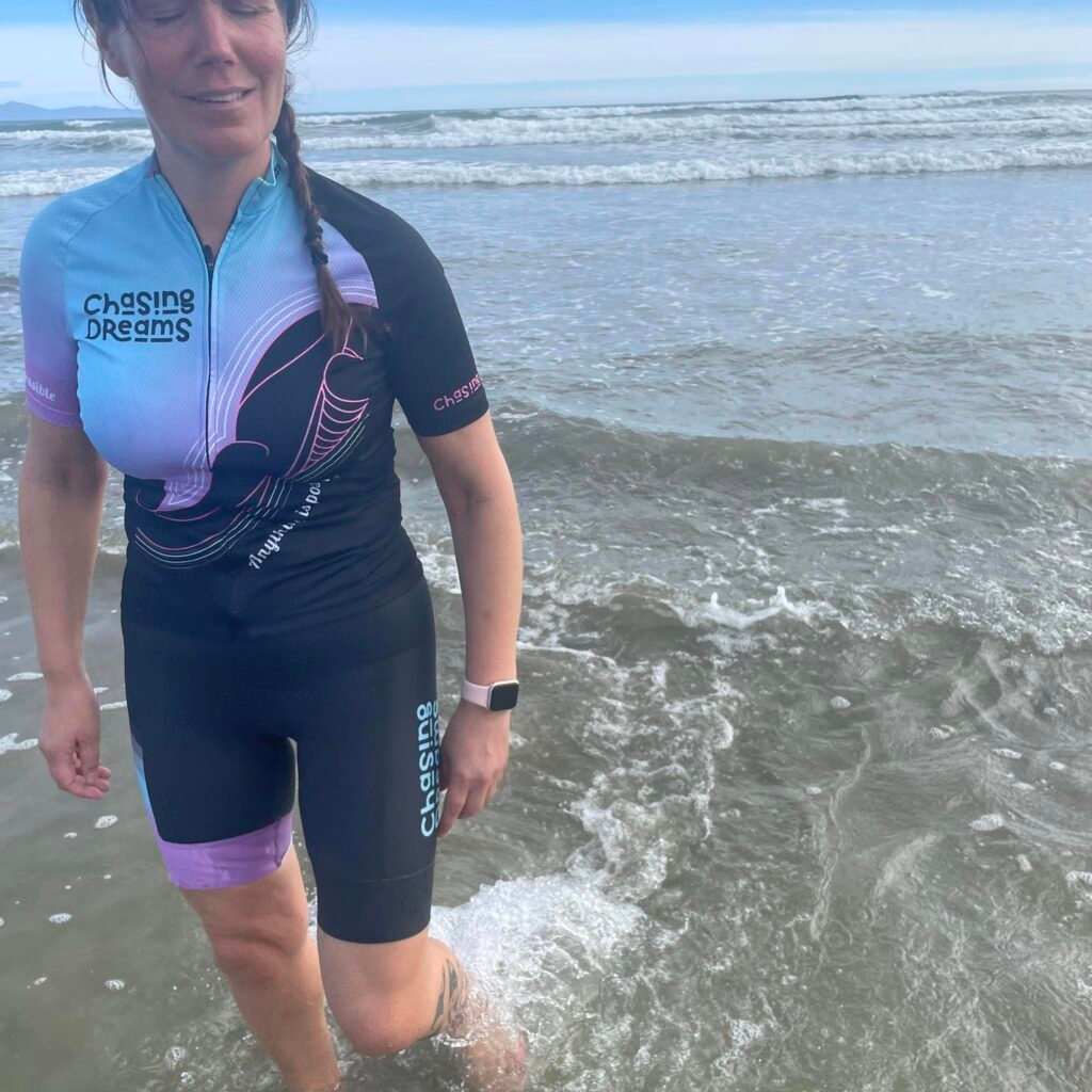 Hannah walking out of the ocean with the beach behind her, wearing her Chasing Dreams cycling kit.