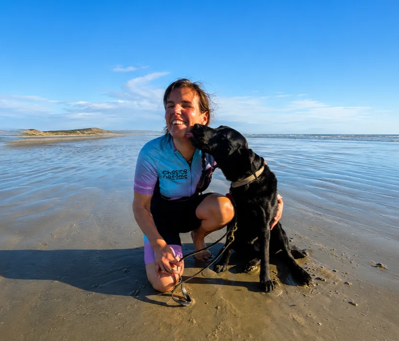 With the beach in the background, Hannah wears her Chasing Dreams cycling kit while her black guide dog Dara licks her face.
